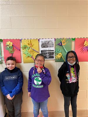 Photo shows three students standing in front of artwork hung on a wall. The student-created work is in the style of A. Thomas